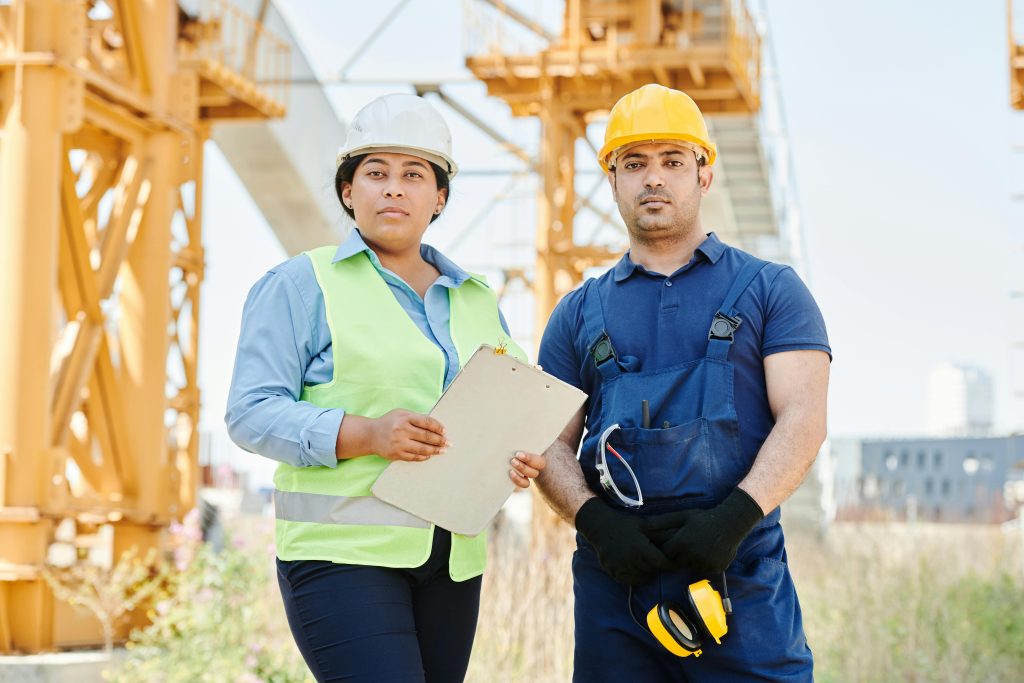 Two engineers at a construction site wearing hard hats and safety vests, standing confidently.